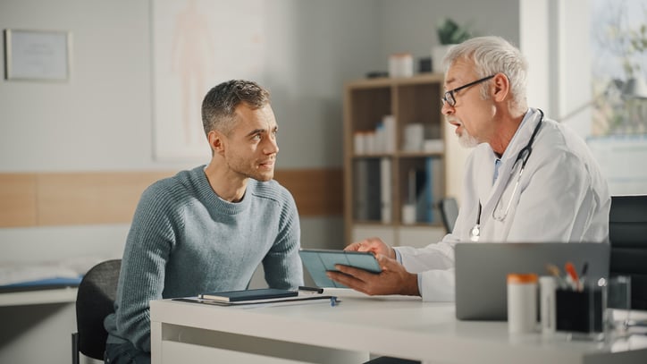 Experienced-Middle-Aged-Family-Doctor-Showing-Analysis-Results-on-Tablet-Computer-to-Male-Patient-During-Consultation-in-a-Health-Clinic.-Physician-Sitting-Behind-a-Desk-in-Hospital-Office.-1315185719_