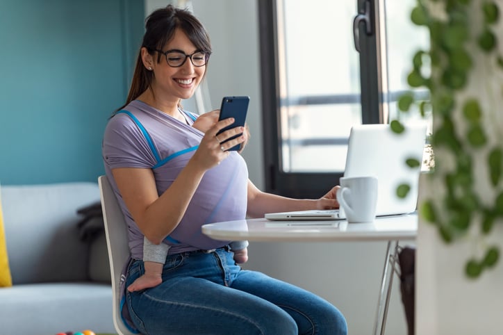 Pretty-young-mother-with-her-baby-in-sling-using-her-mobile-phone-while-working-with-laptop-at-home.-1213213613_6654x4436