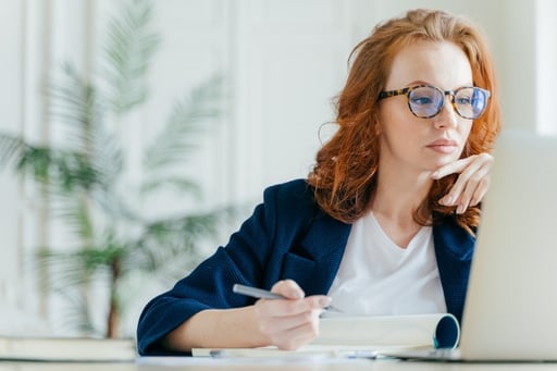 Serious-experienced-female-coach-makes-records-in-notepad,-focused-at-screen-of-laptop-computer,-has-long-red-hair,-wears-transparent-glasses,-watches-webinar-online.-Business-and-job-concept-1139704348_6000x4000-1-1