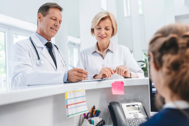 Two-doctors-colleagues-standing-at-reception-desk-talking-and-sharing-information-with-receptionist-in-modern-clinic-1275072738_3867x2579