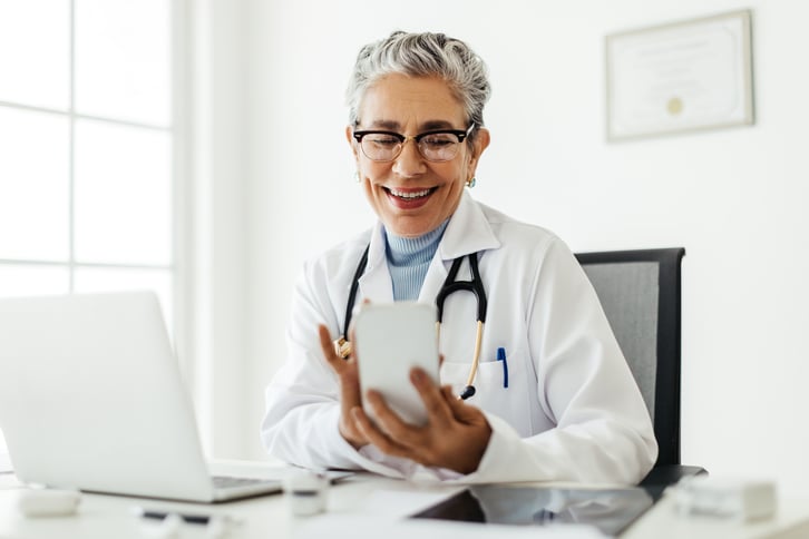 female-european-elderly-doctor-consultation-room-using-her-cellphone-in-her-office-web